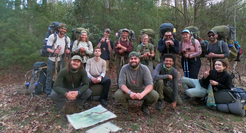 A group of people gathered in a wooded area smile for a group photo. Most of them are wearing backpacks. 